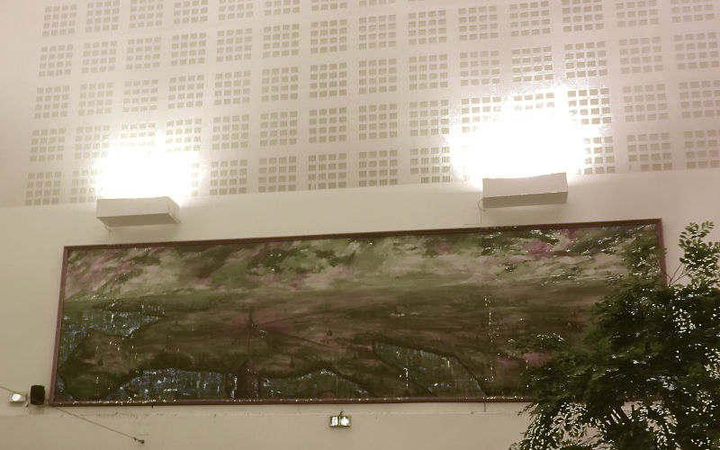 Photo of a train station hall with bright lamps illuminating a geometric wall decoration, while the actual painting (that should be illuminated?) hangs below the lamps and is very dirty and full of pigeon shit. It shows the city of Belfort as center of the world (that is France)